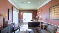 This single-floor space makes a strong visual impression through its rich terracotta-colored accent walls, which are offset by crisp white crown molding and a light ceiling that amplifies the sense of openness. Recessed lighting is distributed evenly throughout the ceiling, supplemented by a decorative pendant chandelier that adds a refined focal point above the central area.

A prominent dark-paneled counter or bar island anchors the middle of the room, featuring a granite or stone surface that adds texture and sophistication. Flanking the counter, bar-height chairs and a bistro-style table are positioned near large windows dressed with white horizontal blinds, allowing natural light to filter softly into the space.

The lounge area to one side includes a comfortable sofa and armchair arranged over a patterned area rug, creating an inviting setting for casual conversation or relaxation. Wall art is displayed in a gallery-style arrangement on the terracotta walls, contributing to the curated, polished atmosphere. A table lamp adds warm ambient lighting near the seating area. Overall, the space balances functional design with an inviting aesthetic.