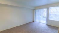 This single-floor space offers a clean, understated layout designed for comfortable everyday living. The main living area is finished with wall-to-wall carpeting in a neutral tone, providing a soft and inviting atmosphere underfoot. Crisp, light-colored walls contribute to an airy, open feel throughout the space. A prominent floor-to-ceiling sliding glass door dominates one wall, dressed with vertical blinds that allow for flexible light control. When open, this feature bathes the interior in abundant natural light and provides a direct visual connection to the outdoors. The overall design is simple and functional, with a neutral palette that serves as a versatile canvas for a variety of furnishing styles. The layout is straightforward and easy to navigate, making efficient use of the available footprint. The combination of soft flooring, bright walls, and generous natural light creates a welcoming ambiance that feels both calm and livable throughout the day.