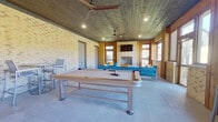 This single-level covered entertainment space combines rustic and contemporary design elements to create a versatile recreational area. The structure is defined by light-toned brick walls and a distinctive wood-plank ceiling that adds warmth and texture throughout. Recessed lighting and multiple ceiling fans ensure the space remains comfortable and well-lit at all times.

A full-size pool table serves as the centerpiece of the main area, positioned on large-format tile flooring that extends throughout the space. Adjacent to the pool table, a seating area features a sectional sofa in vibrant blue with colorful accent pillows, oriented around a brick fireplace. A wall-mounted flat-screen television above the fireplace makes the lounge well-suited for both gaming and media viewing.

Large windows and glass doors along the perimeter allow abundant natural light to fill the space while providing open views of the surrounding neighborhood. The windows also offer direct access to an outdoor area equipped with additional seating. Two interior doors provide access to separate rooms, likely restrooms or utility spaces, flanking the lounge seating area. A bistro-style table and chairs occupy one corner, offering a casual spot for additional seating. The overall ambiance is relaxed, functional, and well-appointed for community or recreational use.