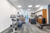 Located at 2382 Kingston Rd in Toronto's Kingston Road corridor, this well-appointed single-floor commercial space offers a thoughtfully designed layout that blends presentation, collaboration, and wellness functions. The interior is finished with a contemporary palette of cool grey walls, patterned carpet tile flooring, and a suspended grid ceiling with recessed LED panel lighting throughout.

The main area serves as a dual-purpose lounge and presentation zone, anchored by two warm wood-paneled feature walls, each housing a wall-mounted flat-screen display. A dark conference-style table surrounded by sleek white chairs provides a formal meeting setup, while a grey sofa and accent chairs create a relaxed lounge atmosphere nearby. A scale architectural model is prominently displayed, suggesting this space functions as a sales or presentation centre. Large floor-to-ceiling glazed entry doors allow ample natural light to pour in and offer clear sightlines to the street.

A dedicated fitness area is accessible through glass-partitioned sliding doors, equipped with a treadmill, elliptical, and stationary bike, catering to active lifestyle needs. The open-plan design ensures seamless flow between zones, while the glass partitions maintain visual connectivity throughout. Overall, the space exudes a professional yet welcoming ambiance, well-suited for client engagement and community amenity use.