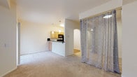This single-floor unit offers a practical, open-concept layout that connects the living area and kitchen in a seamless flow. The spacious living area is finished with neutral wall-to-wall carpeting, providing a warm, understated foundation. A curtained partition on a ceiling-mounted rod creates a flexible division, separating a private sleeping alcove from the main living space. The alcove is carpeted and features a built-in shelf, offering a compact yet functional resting area.

The kitchen is positioned on a raised platform accessible via an arched entryway and is defined by a breakfast bar with dark granite-style countertops. Light wood cabinetry lines the walls and provides ample storage, while black appliances — including a refrigerator, electric range, built-in microwave, and dishwasher — complete the space. Warm hardwood-style flooring runs throughout the kitchen, contrasting with the carpeted living areas. Pendant lighting illuminates both the kitchen and the pass-through bar.

The bathroom features stone-look tile flooring, a light wood vanity with dark countertops, a large mirror, and an open under-vanity design that provides accessible clearance — indicating thoughtful, inclusive design throughout the unit.