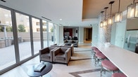 This thoughtfully designed single-floor amenity space in Dallas offers a sophisticated and welcoming environment across several distinct yet connected zones. The lounge area is anchored by plush, taupe upholstered seating arrangements set atop light hardwood-style flooring laid in a herringbone pattern, creating an inviting atmosphere for socializing or relaxing. Floor-to-ceiling glass walls and sliding doors bathe the interior in abundant natural light while providing unobstructed views of a well-appointed outdoor courtyard, which features covered pergola structures, dining tables, and a built-in grilling station with paved hardscaping.

A decorative lattice-style screen partition with a bold geometric pattern adds an artful architectural detail that elegantly defines separate areas without fully enclosing them. The bar area is a standout feature, boasting a long quartz countertop with seating, dark wood cabinetry, a mosaic tile backsplash, stainless steel appliances, a sink, and dual wall-mounted flat-screen televisions. Overhead, a series of bronze lantern-style pendant lights adds warmth and visual interest. The overall ambiance balances modern design sensibility with comfortable, functional elements suited for both casual gatherings and more structured entertaining.