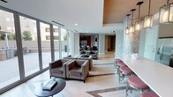 This thoughtfully designed single-floor amenity space in Dallas offers a sophisticated and welcoming environment across several distinct yet connected zones. The lounge area is anchored by plush, taupe upholstered seating arrangements set atop light hardwood-style flooring laid in a herringbone pattern, creating an inviting atmosphere for socializing or relaxing. Floor-to-ceiling glass walls and sliding doors bathe the interior in abundant natural light while providing unobstructed views of a well-appointed outdoor courtyard, which features covered pergola structures, dining tables, and a built-in grilling station with paved hardscaping.

A decorative lattice-style screen partition with a bold geometric pattern adds an artful architectural detail that elegantly defines separate areas without fully enclosing them. The bar area is a standout feature, boasting a long quartz countertop with seating, dark wood cabinetry, a mosaic tile backsplash, stainless steel appliances, a sink, and dual wall-mounted flat-screen televisions. Overhead, a series of bronze lantern-style pendant lights adds warmth and visual interest. The overall ambiance balances modern design sensibility with comfortable, functional elements suited for both casual gatherings and more structured entertaining.