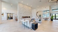 Located at The Reserve in Apopka, Florida, this expansive single-floor clubhouse offers a refined and modern amenity space designed for both relaxation and socializing. The interior is defined by soaring ceilings, large-format light-toned tile flooring, and an abundance of natural light streaming through floor-to-ceiling black-framed windows that frame lush outdoor views.

The centerpiece of the main lounge is a dramatic full-height marble-clad accent wall housing a sleek linear fireplace, creating an impressive focal point for the open seating area. Multiple distinct lounge zones are thoughtfully arranged throughout, featuring contemporary furnishings including plush sectionals, accent chairs, and sculptural coffee tables that contribute to the upscale aesthetic.

A striking geometric pendant chandelier anchors the entrance area, complemented by curated artwork and decorative wall installations. Industrial-style sliding glass barn doors provide flexible separation between spaces, including a dedicated dining and co-working area with its own designer lighting. The game room adds a recreational dimension, complete with a pool table. Tall planters with ornamental grasses punctuate the open floor plan, adding organic texture to the bright, gallery-like atmosphere throughout.