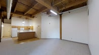 This single-floor space showcases a distinctive rustic-industrial aesthetic defined by its striking exposed wood beam ceiling, which adds warmth and architectural character throughout. The open main area features speckled low-pile carpeting that contrasts with the raw, structural ceiling elements above, including visible wooden joists, support beams, and utility lines that contribute to the space's industrial charm. A central wooden support column anchors the room visually while reinforcing the building's original construction character. Fluorescent tube lighting and hanging pendant fixtures provide ample illumination throughout.

Along one side of the space, a kitchenette area offers wood-toned cabinetry, a countertop with a sink, and what appears to be basic appliances, making the space functional for everyday use. A doorway near the kitchenette leads to an adjacent room, providing a degree of separation within the layout. The walls are finished in clean white, creating a bright contrast against the warm tones of the wood ceiling and cabinetry. Overall, the space delivers a compact yet characterful environment with a blend of utilitarian and rustic design elements.