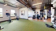 Located at 285 Uptown Blvd in Altamonte Springs, Florida, this fitness center offers a thoughtfully designed, generously sized workout facility spread across a single floor. The space is divided into distinct functional zones, each tailored to a different style of training. An open functional fitness area features turf flooring and is equipped with a multi-purpose rig holding kettlebells, battle ropes, TRX suspension trainers, medicine balls, and a heavy punching bag, providing a versatile space for dynamic workouts. Large mirrors line the walls, amplifying the sense of space and supporting form-conscious training. A dedicated cardio zone includes treadmills, a rowing machine, and a stationary bike, positioned near windows that bring in natural light and outdoor views. The strength training area is furnished with cable machines, selectorized equipment, and a full dumbbell rack paired with flat benches, catering to a wide range of resistance training needs. Hardwood-style flooring transitions seamlessly between zones, complemented by an industrial-style exposed ceiling with visible ductwork and sleek linear lighting that gives the facility a modern, urban aesthetic. Tasteful artwork on the walls adds a refined touch to the energetic environment, making this fitness center both functional and visually inviting.