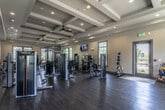 This single-floor fitness center offers a comprehensive workout environment designed with both functionality and aesthetics in mind. The space is anchored by a striking coffered ceiling accented with recessed lighting and ceiling fans, lending the room a refined, upscale character that sets it apart from a typical gym setting. Dark hardwood-style flooring runs throughout, providing a polished foundation that complements the neutral warm-toned walls.

The facility is outfitted with a full suite of Matrix-brand strength training machines, strategically arranged to allow comfortable circulation between stations. A dedicated free weight area features a tiered dumbbell rack alongside floor mirrors that enhance both the sense of space and utility for form-focused training. Cardio equipment, including multiple treadmills and stair climbers with personal monitors, is positioned near large windows that frame outdoor views and flood the space with natural light.

Additional functional training equipment is also present, including medicine balls and kettlebell racks, catering to a variety of workout routines. Wall-mounted flat-screen televisions provide entertainment throughout the space. Two glass-panel exterior doors offer direct outdoor access, contributing to an open and airy atmosphere. Overall, the fitness center strikes a balance between a well-appointed design and a practical, fully equipped training environment.