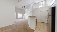 This two-story home is defined by its dramatic vaulted ceilings in the main living area, which create a sense of expansive vertical space and are complemented by a ceiling fan and recessed lighting. The open-concept layout on the ground floor connects the living area—finished with plush neutral carpet—to the entry and kitchen zones, which feature light tile flooring. A white-railed staircase ascends to a second-floor landing with a balcony-style overlook, adding architectural interest and visual depth to the main level.

The kitchen is equipped with white cabinetry fitted with brushed nickel hardware, granite-style countertops, a white subway tile backsplash, and a stainless electric range. A peninsula with a sink provides additional workspace and separates the kitchen from the living area. Upstairs, the bedrooms are carpeted and receive natural light through windows with outdoor views of mature trees. Each room includes a ceiling fan. The bathrooms feature tile flooring, white cabinetry with granite-style countertops, and large mirrors. One bathroom includes a walk-in closet, while another offers a tub-shower combination. The overall aesthetic is clean and neutral throughout.