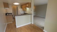 This single-floor residence offers a practical and comfortable layout highlighted by an open-concept design that connects the kitchen and living area. The kitchen is well-appointed with warm oak cabinetry, dark granite-style countertops, and a peninsula that doubles as a breakfast bar, providing additional workspace and casual seating. A white refrigerator and under-cabinet lighting add functional touches to the cooking space. The kitchen transitions naturally into the living area, which is distinguished by soft, neutral-toned carpeting that creates a cozy and inviting atmosphere. The flooring shifts from warm hardwood in the kitchen and dining areas to carpet in the living space, clearly delineating each zone while maintaining a cohesive aesthetic throughout. Light-toned walls in a soft cream hue reflect the overhead lighting and contribute to a bright, airy feel. A flush-mount ceiling light in the kitchen and additional lighting visible in the adjoining room ensure the space is well-lit. The open sightlines throughout the main living areas make the space feel generous and connected, ideal for everyday living and casual entertaining. Clean white baseboards and trim provide a crisp finish throughout.