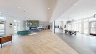 This expansive single-floor amenity center offers a thoughtfully designed layout that seamlessly blends social, recreational, and wellness spaces. The main lounge area serves as the heart of the facility, featuring a striking stone fireplace framed by a teal accent wall, a mounted flat-screen TV, and curated botanical wall art. Light-toned herringbone hardwood flooring runs throughout, adding warmth and visual continuity. Abundant natural light pours in through large floor-to-ceiling windows and glass entry doors, creating an open, airy atmosphere. Sculptural pendant lighting fixtures add a contemporary design accent overhead.

Adjacent to the lounge is a versatile event and co-working area complete with a long communal table, lounge seating, and a well-appointed kitchenette outfitted with navy cabinetry, gold hardware, white countertops, open wood shelving, and a textured fluted accent wall. A game room with a ping-pong table and additional seating rounds out the social spaces.

The fitness center is equipped with cardio machines, cable and strength equipment, free weights, and full-length mirrors, all bathed in natural light from surrounding windows. A dedicated multi-purpose studio space with high ceilings, green accent walls, and patterned geometric flooring completes the wellness amenities, alongside a water station and private restrooms.