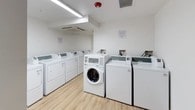 This single-floor shared laundry facility is designed with functionality and cleanliness in mind. The space is finished with crisp white walls and durable light wood-look luxury vinyl plank flooring, creating a bright and hygienic environment that is easy to maintain.

The main laundry area is the centerpiece of the space, housing a generous lineup of commercial-grade top-loading washers and dryers arranged in parallel rows for efficient use. A front-loading washer is also present, offering additional flexibility for residents. The machines are neatly aligned and well-maintained, reflecting a modern, organized approach to shared laundry amenities. Overhead fluorescent strip lighting ensures the room is well-lit at all times, and a wall-mounted HVAC unit provides climate control for year-round comfort.

Safety features are prominently integrated throughout, including visible fire sprinkler heads and smoke detectors mounted on the ceiling. Informational signage is posted on the walls to guide proper use of the equipment.

Adjacent utility or transition rooms feature the same consistent flooring and white wall finishes, with access control panels and solid-core doors fitted with lever hardware. The overall design is minimal and practical, prioritizing ease of use, cleanliness, and safety for all occupants.