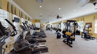 This fitness center offers a comprehensive workout environment thoughtfully designed to accommodate a wide range of exercise needs. The space is finished with warm yellow walls, crown molding, and neutral carpeted flooring, lending it an inviting, upscale character that sets it apart from a typical gym setting.

The cardio zone is prominently featured along one wall, housing a row of elliptical trainers and treadmills, each equipped with individual monitor screens for entertainment during workouts. Ceiling fans with decorative light fixtures provide comfortable air circulation throughout. Large windows dressed with valances line the walls, allowing generous natural light to fill the space while offering outdoor views, and a full-length mirror along one wall visually expands the room.

The strength training area includes a well-stocked dumbbell rack alongside multiple cable and resistance machines with tan upholstered seating, providing options for both isolated and compound movements. A pull-up and dip station adds further versatility. Access to the facility is provided through elegant French doors, and recessed lighting throughout ensures consistent, even illumination. The overall ambiance is polished and functional, balancing aesthetic appeal with a full suite of fitness amenities.