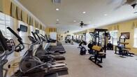 This fitness center offers a comprehensive workout environment thoughtfully designed to accommodate a wide range of exercise needs. The space is finished with warm yellow walls, crown molding, and neutral carpeted flooring, lending it an inviting, upscale character that sets it apart from a typical gym setting.

The cardio zone is prominently featured along one wall, housing a row of elliptical trainers and treadmills, each equipped with individual monitor screens for entertainment during workouts. Ceiling fans with decorative light fixtures provide comfortable air circulation throughout. Large windows dressed with valances line the walls, allowing generous natural light to fill the space while offering outdoor views, and a full-length mirror along one wall visually expands the room.

The strength training area includes a well-stocked dumbbell rack alongside multiple cable and resistance machines with tan upholstered seating, providing options for both isolated and compound movements. A pull-up and dip station adds further versatility. Access to the facility is provided through elegant French doors, and recessed lighting throughout ensures consistent, even illumination. The overall ambiance is polished and functional, balancing aesthetic appeal with a full suite of fitness amenities.