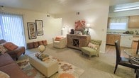 This single-floor space showcases a thoughtfully designed living area that balances comfort and style. The main living room is generously proportioned, featuring neutral carpet flooring complemented by a floral-patterned area rug that adds a vibrant focal point to the room. Warm, cream-toned walls provide a versatile backdrop for the eclectic mix of furnishings throughout.

The living area is furnished with a variety of seating options, including a tufted ottoman, a sage green accent chair with floral cushioning, and a swivel armchair in neutral tones. A wooden sideboard with wine storage serves as a functional centerpiece, topped with decorative items including a table lamp and floral artwork, adding warmth and character to the space.

Decorative wall art, including a framed map print, adds visual interest, while floor and table lamps provide layered ambient lighting. Visible HVAC vents are integrated into the ceiling and walls for climate control. An adjacent area, partially visible in the background, hints at additional rooms, suggesting a practical and connected floor plan. Overall, the space projects a warm, lived-in ambiance.