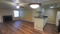 This single-floor space offers a well-designed open-concept layout that seamlessly connects the living area and kitchen, creating a cohesive and functional environment. Rich, dark hardwood flooring runs throughout, adding warmth and character to the interior. The living area is bright and airy, benefiting from natural light that streams in through blinded windows, and is complemented by a ceiling fan that enhances comfort year-round. Crown molding along the upper walls adds a refined architectural detail, while the textured ceiling provides additional visual depth. The kitchen is thoughtfully appointed with white cabinetry offering generous storage, dark granite countertops, and a peninsula that doubles as a breakfast bar — ideal for casual dining or additional prep space. A striking pendant light fixture hangs above the peninsula, serving as a focal point and providing warm task lighting. The kitchen also features a built-in cooktop and a sink integrated into the peninsula, maximizing efficiency. The neutral wall tones throughout create a versatile backdrop, allowing the natural materials and finishes to take center stage. Overall, the space strikes a balance between practicality and style, making it a comfortable and inviting environment.