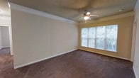 This single-floor space offers a comfortable and well-appointed living area designed with functional elegance. The main living room is generously sized, featuring plush wall-to-wall carpeting that adds warmth underfoot. Crisp white crown molding frames the room with a clean, classic finish, while a textured ceiling adds subtle visual interest overhead. A ceiling fan with an integrated light fixture is centrally positioned, providing both air circulation and ambient lighting throughout the space. A large multi-pane window along one wall floods the room with natural light and offers pleasant views of the surrounding greenery, creating a bright and inviting atmosphere. Neutral-toned walls complement the overall palette, lending the space a versatile and timeless quality that easily accommodates a variety of interior styles. The layout is open and uncluttered, maximizing usable floor space and allowing for flexible furniture arrangements. White baseboards provide a clean contrast against the darker carpeting, reinforcing the tidy, well-maintained character of the space. Overall, this home offers a calm and practical living environment with thoughtful architectural details throughout.