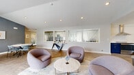 This thoughtfully designed single-floor space functions as a communal gathering area, blending modern aesthetics with practical amenities across several distinct zones. The entrance corridor sets the tone with herringbone-patterned hardwood flooring, a bold teal textured accent wall, and frosted glass-paneled doors that introduce an air of contemporary sophistication.

The central lounge area is anchored by a striking marble-surround fireplace with a wall-mounted television, flanked by plush rounded chairs in muted mauve tones arranged around a marble-top coffee table. French doors provide direct access to an outdoor patio, filling the space with natural light and outdoor views. Recessed lighting and vaulted ceilings enhance the open, airy feel.

Adjacent to the lounge, a dedicated dining area features marble-top round tables paired with upholstered side chairs, set against textured gray accent walls adorned with framed artwork. A water cooler and additional seating make it suitable for meetings or casual gatherings.

The fully equipped kitchen showcases bold navy cabinetry, white quartz countertops, open wood shelving, stainless steel appliances including a range, dishwasher, microwave, and beverage refrigerator, all illuminated by a modern circular light fixture. Throughout, herringbone flooring ties the spaces together cohesively.