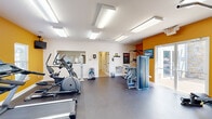 Located at 3300 Stadium Dr in Phenix City, Alabama, this fitness center offers a well-organized, single-room workout facility designed to meet a range of exercise needs. The space is defined by crisp white walls accented by a bold orange feature wall, creating an energetic and motivating atmosphere. Durable rubber flooring in a speckled gray finish covers the entire area, providing both safety and comfort during workouts.

The room is equipped with a variety of cardio machines, including multiple treadmills, elliptical trainers, and a stationary bike, all arranged to maximize open floor space. For strength training, the center includes a multi-function cable/weight machine, a vertical knee raise and pull-up tower, and a dumbbell rack with free weights. A wall-mounted television and framed artwork add to the functionality and ambiance of the space.

Bright recessed panel lighting across the ceiling ensures the room is evenly and thoroughly illuminated. A pass-through window connects to an adjacent office or service area, and a doorway leads to additional rooms. A sliding glass door on the orange accent wall fills the corner with natural light and provides direct access to the outdoor area. A wall-mounted water fountain offers convenient hydration. Posted fitness center rules and instructional charts reflect a well-managed facility.