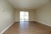 This single-floor unit offers a practical and cohesive layout connecting a living area, kitchen, bedroom, and bathroom. The living room is generously proportioned, featuring dark wood-look flooring, neutral warm-toned walls, and a textured ceiling. A sliding glass door fills the space with natural light and provides views of the surrounding greenery, while vertical blinds offer privacy control. An open passage leads directly into the galley-style kitchen, which is outfitted with dark espresso cabinetry both above and below the counters, granite-look countertops, a double-basin stainless steel sink, and a tile backsplash accent. Black appliances, including a refrigerator and electric range, complete the functional cooking space. The bedroom continues the warm aesthetic with plush carpeting, a ceiling light fixture, a vertical-blind window admitting natural light, and ample wall space. The bathroom features wood-look flooring, a granite-topped vanity, and a full tub-shower combination enclosed by large-format neutral tile. Throughout the unit, consistent design choices in flooring, cabinetry finishes, and warm wall tones create a unified and cohesive ambiance.