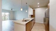 This single-floor residence showcases a thoughtfully designed open-concept layout with a consistent palette of warm wood tones, light wood-style flooring, and neutral walls that create a cohesive, contemporary aesthetic throughout. The kitchen serves as a central hub of the living area, anchored by a peninsula island topped with granite-style countertops and an undermount stainless steel sink. Pendant lighting illuminates the workspace, while a full suite of stainless steel appliances — including a refrigerator, range, microwave, and dishwasher — provides modern functionality. Upper white cabinetry is complemented by warm wood lower cabinets, adding visual depth. The living area flows seamlessly from the kitchen, featuring light hardwood-style flooring and a soft, airy color scheme. Both bedrooms feature plush wall-to-wall carpeting and windows fitted with blinds that invite natural light. One bedroom includes a stylish sliding barn door leading directly into an en-suite bathroom, while another bedroom connects to a full bathroom with walk-in closet access. Each bathroom is outfitted with a warm wood vanity, white countertop, large mirror, and light hardwood-style flooring, maintaining the space's clean, modern character throughout.