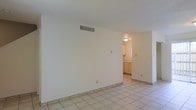 This two-floor space presents a clean, minimalist interior defined by light-toned ceramic tile flooring and smooth white walls throughout. The main living area is open and simply appointed, offering a blank canvas well-suited for a variety of furniture arrangements. A ceiling-mounted HVAC vent ensures year-round climate control, while flush ceiling lighting provides consistent illumination across the space.

A wide doorway connects the living area to the kitchen, which is visible from the main room and features light wood cabinetry, matching tile flooring, and its own ceiling fixture. The cabinetry offers practical storage, and the layout appears functional and efficiently organized.

The overall aesthetic is neutral and understated, with white walls and pale flooring contributing to a bright, airy atmosphere throughout. The consistent use of tile flooring across both the living and kitchen areas creates visual continuity. With two floors, the space offers additional room beyond what is immediately visible, providing versatility in how the layout can be utilized across different levels.