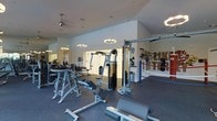 This single-level fitness center offers a thoughtfully designed workout space equipped with a comprehensive range of exercise machines and training stations. The room features vaulted ceilings that lend an open, airy feel, complemented by a combination of recessed lighting and stylish pendant light fixtures that ensure the space is well-illuminated throughout. The flooring consists of durable rubber gym matting, providing safety and comfort during workouts. A large mirror along one wall enhances the sense of space while allowing users to monitor their form. The facility includes multiple strength-training machines, free weight stations, a cable machine, and what appears to be a boxing or functional training area, complete with a heavy bag and resistance equipment. A motivational phrase is displayed prominently on the wall, adding an energetic character to the environment. A ceiling fan contributes to air circulation, keeping the space comfortable during intense sessions. The clean, white walls and neutral tones create a focused, professional atmosphere well-suited for fitness enthusiasts of all levels.
