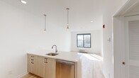 This thoughtfully designed 1-bedroom, 1-bathroom unit offers a clean, modern aesthetic defined by crisp white walls, light hardwood flooring, and an abundance of natural light. The open-concept layout seamlessly connects the kitchen and living area, creating a bright and functional living space. The kitchen is well-appointed with stainless steel appliances, including a French door refrigerator, electric range, over-the-range microwave, and dishwasher. Light wood cabinetry with matte black hardware pairs with a white quartz countertop and peninsula, which is illuminated by two stylish glass pendant lights — ideal for casual dining or meal prep. A light gray subway tile backsplash adds a refined touch. The living area benefits from a large black-framed window that frames an urban view and allows generous sunlight to fill the space. The bedroom is a comfortable, well-proportioned retreat featuring a double-door closet, hardwood floors, and its own window with an outdoor view. The bathroom features large-format floor tiles, a floating wood vanity, a rounded rectangular mirror, and a well-lit vanity bar, delivering a clean and contemporary finish throughout. A hallway connects all areas efficiently, completing this compact yet fully functional layout.