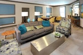 This single-floor community lounge is thoughtfully designed to accommodate a variety of social and recreational activities within one open, flowing space. The layout is anchored by a full-size pool table positioned beneath a stylish pendant light fixture, with large windows nearby offering natural light and views of surrounding greenery. Adjacent to the billiards area is a comfortable lounge zone featuring plush neutral sofas accented with teal throw pillows, a wooden console table with decorative lamps, and an accent chair with a bold geometric pattern.

A dedicated game and card table area occupies one corner of the room, furnished with a round dark-wood table and yellow upholstered chairs on casters, complemented by colorful abstract artwork on the walls. Nearby, a wet bar with a dark countertop and bar-height seating provides a convenient refreshment station. A wall-mounted flat-screen television and media console anchor another seating arrangement, creating a relaxed viewing area.

Throughout the space, patterned low-pile carpeting, recessed lighting, and a neutral color palette with teal and gray accents contribute to a cohesive, contemporary ambiance. Multiple windows ensure consistent natural light, while the open floor plan allows for easy movement between activity zones.