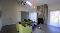 This well-appointed single-floor space offers a sophisticated and inviting atmosphere, thoughtfully designed with contemporary finishes and curated décor. The lounge area serves as the focal point of the space, anchored by a floor-to-ceiling tiled fireplace wall in warm grey tones, accented by a decorative sunburst mirror that adds artistic flair. Vaulted ceilings lend an airy, expansive feel to the interior, enhanced by a skylight that draws in natural light and a sleek ceiling fan for year-round comfort. A striking geometric chandelier hangs overhead, serving as a bold design statement that complements the modern aesthetic. Seating is arranged around a dark coffee table, with vibrant chartreuse upholstered chairs and coordinating striped accent pillows providing a lively pop of color against the neutral backdrop. A set of sliding barn-style doors in a muted grey finish adds a functional yet stylish architectural detail, offering flexible space division. Vertical blinds along the windows allow for adjustable natural light. The overall ambiance is modern and welcoming, balancing clean lines and neutral tones with carefully chosen accents that create a warm, refined environment suited for relaxation or social gatherings.