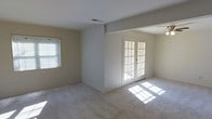 This single-story space presents a clean and straightforward layout characterized by neutral tones and practical finishes. The main living area is finished with wall-to-wall carpeting in a light beige tone, contributing to a warm and understated atmosphere. Walls are painted in a soft, neutral shade that complements the overall palette and enhances the sense of openness throughout the space.

A prominent architectural feature is the set of white-framed French doors, fitted with multi-pane glass panels that allow generous natural light to pour into the interior. The resulting sunlight creates bright patterns across the carpeted floor, adding a lively quality to the otherwise understated room. A standard window fitted with horizontal blinds on the adjacent wall provides an additional source of natural light and ventilation.

A partial wall or column detail creates a subtle division within the open layout, offering a sense of structure without fully enclosing the space. Ceiling-mounted HVAC vents are visible, indicating a central air system, and a ceiling fan is partially visible in the adjoining area, supporting year-round comfort. Overall, the space offers a functional, move-in-ready environment with a calm, neutral aesthetic.