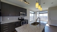 Located at 900 Luxor Lane in Norristown, Pennsylvania, this single-floor residence offers a thoughtfully designed living space with a distinctly modern aesthetic. The kitchen serves as a standout centerpiece, showcasing rich dark cabinetry that contrasts elegantly against light granite countertops with natural veining. Stainless steel appliances, including an electric range with oven and an over-the-range microwave, lend the kitchen a clean, contemporary feel. A large kitchen island anchors the space and features a deep undermount sink with a sleek high-arc faucet, providing both style and functionality. Overhead, a trio of warm pendant lights adds an inviting ambiance while track lighting illuminates the cooking area with precision. Expansive corner windows flood the kitchen with natural light and frame sweeping views of the surrounding Norristown landscape, giving the space an open, airy quality. The windows extend nearly floor to ceiling, maximizing the connection to the outdoors and creating a bright, welcoming atmosphere throughout. The overall design of the space balances practicality with refined style, making it an ideal setting for everyday living and entertaining alike.