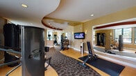 Located at 399 Main Ave in Norwalk, Connecticut, this well-appointed fitness center offers a comprehensive workout environment designed for both cardio and strength training. The space is defined by a striking curved ceiling feature with a bold terra cotta accent band, which adds architectural character and guides the eye throughout the room. Flooring transitions seamlessly between light hardwood and a patterned carpet runner, delineating distinct workout zones.

The cardio section is generously equipped with treadmills, elliptical trainers, spinning bikes, stair climbers, and recumbent bikes, ensuring a wide variety of aerobic training options. A wall-mounted flat-screen television keeps users entertained during workouts. The strength training area features Cybex cable machines, multi-station weight systems, a full dumbbell rack, benches, and additional resistance equipment, providing thorough coverage for full-body conditioning.

Large windows along one wall flood the space with natural light and frame views of the surrounding greenery, creating an energizing atmosphere. Recessed and track lighting supplement the natural light throughout. Mirror panels in the strength area enhance the sense of openness. The overall ambiance is polished and professional, reflecting a facility designed to support a complete and varied fitness regimen.