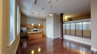 This single-floor space offers an open-concept layout that seamlessly connects the kitchen and living areas, creating a cohesive and inviting environment. The kitchen is well-appointed with warm wood cabinetry, granite countertops, stainless steel appliances including a refrigerator, microwave, and range, and a central island that provides additional prep and gathering space. Pendant lighting hangs above the island, adding a stylish focal point to the room. Throughout the space, rich, dark-toned hardwood flooring adds warmth and a polished aesthetic, reflecting the natural and artificial light beautifully. The ceilings rise generously overhead, enhancing the sense of openness, and are equipped with a ceiling fan for added comfort and track lighting for functional illumination. To the right, a glass partition wall with burgundy accent columns defines a distinct zone within the space, allowing light to pass through while providing visual separation. The neutral gray wall tones provide a versatile backdrop that complements the warm wood finishes. Overall, the space balances modern design elements with warm, inviting materials, resulting in a functional yet stylish interior.