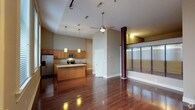 This single-floor space offers an open-concept layout that seamlessly connects the kitchen and living areas, creating a cohesive and inviting environment. The kitchen is well-appointed with warm wood cabinetry, granite countertops, stainless steel appliances including a refrigerator, microwave, and range, and a central island that provides additional prep and gathering space. Pendant lighting hangs above the island, adding a stylish focal point to the room. Throughout the space, rich, dark-toned hardwood flooring adds warmth and a polished aesthetic, reflecting the natural and artificial light beautifully. The ceilings rise generously overhead, enhancing the sense of openness, and are equipped with a ceiling fan for added comfort and track lighting for functional illumination. To the right, a glass partition wall with burgundy accent columns defines a distinct zone within the space, allowing light to pass through while providing visual separation. The neutral gray wall tones provide a versatile backdrop that complements the warm wood finishes. Overall, the space balances modern design elements with warm, inviting materials, resulting in a functional yet stylish interior.