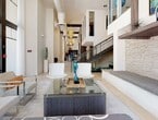 This expansive, single-floor communal space showcases a striking contemporary design defined by soaring double-height ceilings and an open, flowing layout that connects multiple distinct zones seamlessly. The main lounge area draws immediate attention with its dramatic vertical scale, featuring sculptural pendant lighting, crisp white walls, and large floor-to-ceiling windows dressed in sheer curtains that flood the interior with natural light and offer views of the surrounding greenery outside.

Throughout the space, carefully curated artwork and decorative sculptures add personality and visual interest, complementing the refined neutral palette of light tile flooring and textured stone accent walls. Seating areas are thoughtfully arranged, offering a mix of sleek modern chairs, plush leather sofas with warm accent cushions, and glass-topped coffee tables — creating inviting settings for both casual conversation and focused gatherings.

A dedicated lounge section features a wall-mounted flat-screen television, stone-clad accent walls with warm underlighting, and rich brown leather seating arranged around a central coffee table. The kitchenette is well-appointed with warm wood cabinetry, light stone countertops, a blue glass tile backsplash, under-counter refrigeration, and a large communal dining island illuminated by colorful pendant lights. A package hub station adds a practical modern convenience. A staircase with sleek metal railings connects to an upper mezzanine level, adding architectural depth to this beautifully designed space.