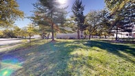 This outdoor dog park offers a generously sized, ground-level green space designed for pets and their owners. The area is defined by a sturdy chain-link fence perimeter, with a gated entry point providing controlled access via a paved walkway. The grounds are covered in well-maintained grass, offering ample open space for pets to roam freely. Several mature trees — including tall evergreen pines and broad deciduous trees — are scattered throughout the park, providing natural shade across much of the area during sunny days. The canopy coverage creates a comfortable environment for both animals and visitors in warmer weather. Benches are positioned within the enclosure, giving owners a place to sit and supervise their pets. The surrounding landscape is neatly kept, with mulched bases around the tree trunks. The park is situated adjacent to a multi-unit residential building, making it a convenient amenity for nearby residents. The open sky above and the natural greenery throughout give the space a pleasant, park-like ambiance. The combination of secure fencing, shaded areas, seating, and open lawn makes this a functional and welcoming outdoor space for pet owners and their companions.