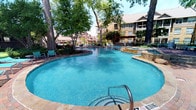 This resort-style outdoor amenity area is designed to offer a comprehensive recreational experience in an open-air setting. At its heart is a generously sized freeform swimming pool with clear blue water, natural stone accents, and multiple entry points, including wide steps that lead gracefully into the water. Mature trees provide natural shade throughout the space, creating a lush, park-like atmosphere under open skies.

A covered outdoor kitchen and bar area anchors one end of the pool deck, featuring granite countertops and pergola-style overhead structure — ideal for gatherings and entertaining. Adjacent to this is a spacious elevated deck finished in rich red-toned wood, furnished with vibrant teal lounge chairs and anchored by a striking stone fireplace, offering a comfortable outdoor living area for relaxation.

Brick-paved walkways wind throughout the grounds, connecting the various amenity zones. A dedicated putting green with well-manicured turf and multiple holes adds a recreational touch, bordered by stone retaining walls and ornamental landscaping. Decorative lamp posts and lush greenery enhance the overall ambiance, while the surrounding architecture — featuring stone facades, pitched roofs, and balconies — frames the entire outdoor space with a cohesive, upscale aesthetic.
