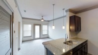 This single-floor residence offers a thoughtfully designed open-concept layout that seamlessly connects the kitchen and living areas. The kitchen is appointed with rich dark cabinetry, granite countertops, and a stainless steel double-basin sink. A trio of elegant pendant lights hangs above the kitchen island, providing both functional task lighting and a refined aesthetic touch.

The living area is generously proportioned, featuring plush carpeting and a ceiling fan that contributes to a comfortable and inviting atmosphere. Natural light fills the space through a set of large windows and a glass-paneled door that opens to an outdoor patio area, framed with white horizontal blinds for adjustable privacy and light control. The neutral, light-toned walls and white textured ceiling complement the overall clean and contemporary design.

Architectural details such as crown molding along the upper walls add a subtle layer of sophistication throughout. The open flow between the kitchen and living space makes the interior feel expansive and well-connected, ideal for both everyday living and entertaining. The overall ambiance is bright, modern, and welcoming, with a cohesive palette of neutral tones and quality finishes throughout.