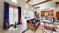 This generously sized single-floor living space is defined by its striking architectural character and thoughtfully curated interior design. The room commands immediate attention with its soaring ceilings, accented by prominent decorative wood beams that add warmth and rustic contrast to the crisp, neutral walls. Large clerestory windows positioned high along the walls flood the space with natural light throughout the day, complementing the oversized sliding glass doors that open directly to an outdoor patio area, creating a seamless indoor-outdoor flow.

The main living area features large-format cream ceramic tile flooring and is anchored by a stylish area rug with bold geometric detailing in warm earth tones. A leather sofa with colorful accent pillows, upholstered armchairs, and a coordinating seating arrangement give the space a welcoming, layered feel. Floor-length drapes in deep chocolate brown with vibrant red paisley accents frame the windows and add a dramatic touch.

A distinctly designed bar or lounge area is set apart by a bold terracotta orange accent wall and a circular tray ceiling with recessed and pendant lighting, adding an upscale, intimate atmosphere. French doors with multi-pane glass panels provide a secondary entrance and allow additional light to pass through, further enhancing the open, airy ambiance of this well-designed space.