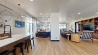 This thoughtfully designed single-floor communal space blends contemporary aesthetics with functional versatility. Light hardwood flooring runs throughout, unifying the various zones and contributing to an airy, welcoming atmosphere. At the heart of the space is a generous bar-height island finished in white marble veining, complemented by warm wood cabinetry with fluted detailing and a row of sleek black bar stools. Dramatic circular pendant lights hang above, adding an artistic focal point. A striking geometric black lattice screen serves as a sculptural room divider, separating a casual lounge area — anchored by a deep navy sofa and accent chairs — from the rest of the open floor plan. A secondary lounge zone features curved mustard-toned armchairs, small accent tables, and floor-to-ceiling windows that flood the space with natural light and offer expansive outdoor views, including a notable stadium sign visible from the exterior glazing. Built-in wood shelving with integrated media display provides a relaxed reading and entertainment nook. A glass-walled private conference or meeting room with a round black dining table and pendant lighting offers a more intimate, enclosed setting. Throughout, curated artwork, decorative objects, and warm neutral tones combine to create a polished yet inviting communal environment.