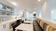 Located at 22121 Clarendon Street in Los Angeles, this single-floor WiFi Room is a thoughtfully designed communal amenity space that blends functionality with modern aesthetics. The layout seamlessly connects several distinct zones, each serving a unique purpose for residents or guests.

The kitchenette area is outfitted with crisp white cabinetry, open display shelving, a penny-tile backsplash, a built-in beverage tap, under-counter refrigeration, and a microwave — all set against a clean white countertop. Adjacent to the kitchenette, a dining area features marble-topped tables paired with sleek black chairs and a generously sized banquette upholstered in a bold multicolor striped fabric, adding a lively accent to the otherwise neutral palette.

Sheer floor-to-ceiling curtains line the exterior-facing walls, softening natural light while maintaining an open, airy feel throughout. A tall potted dracaena plant adds a natural, organic element to the lounge area, which also features a wall-mounted flat-screen TV and minimalist bar-height seating along a floating ledge.

The game room offers a full-size ping pong table and a shuffleboard table, set beneath recessed lighting and framed by a vibrant large-scale abstract artwork. Light wood flooring flows consistently throughout, tying all areas together into one cohesive, contemporary space designed for both productivity and recreation.