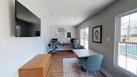 This thoughtfully designed single-floor workspace offers a clean, contemporary environment ideal for focused work and collaboration. The room features warm hardwood-style flooring complemented by light gray walls, creating a bright and professional atmosphere throughout.

A built-in wooden desk shelf runs along the back wall, accommodating two full workstations complete with monitors and ergonomic Herman Miller chairs — a setup that speaks to a commitment to comfort and productivity. Motivational wall art and decorative accents lend the space a modern, creative personality without sacrificing professionalism.

Adjacent to the workstations, a compact meeting area includes a square table and a pair of teal upholstered chairs, providing a comfortable spot for small group discussions or one-on-one meetings. A large window along the side wall floods the room with natural light, enhancing the open and welcoming feel of the space.

A wall-mounted flat-screen television is positioned prominently for presentations or collaborative viewing. A wooden storage credenza anchors one side of the room, offering practical storage while contributing to the warm, cohesive aesthetic.

Overall, this workspace balances function and style, making it a productive and inviting environment for both individual work and small team collaboration.