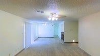 This single-floor space features an open-concept layout that combines the living and dining areas into one expansive, flowing zone. The interior is finished with neutral-toned carpeting throughout, providing a clean and versatile foundation for furnishing. Two ceiling fans with integrated light fixtures are positioned across the living area, ensuring consistent airflow and ambient lighting throughout the space.

The textured popcorn ceiling adds a classic residential character overhead, while the smooth, light-colored walls contribute to a bright and airy atmosphere. Vertical blinds on the left wall cover what appears to be a large window or sliding door, allowing for adjustable natural light and potential outdoor access.

Toward the rear of the space, a partial view of the kitchen is visible, revealing dark cabinetry and a countertop transition. A doorway nearby provides access to additional rooms within the unit. The open sightlines between the living area and kitchen create a connected, functional flow well-suited for everyday living. Overall, the space presents a straightforward, practical interior with key comfort features throughout.