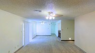 This single-floor space features an open-concept layout that combines the living and dining areas into one expansive, flowing zone. The interior is finished with neutral-toned carpeting throughout, providing a clean and versatile foundation for furnishing. Two ceiling fans with integrated light fixtures are positioned across the living area, ensuring consistent airflow and ambient lighting throughout the space.

The textured popcorn ceiling adds a classic residential character overhead, while the smooth, light-colored walls contribute to a bright and airy atmosphere. Vertical blinds on the left wall cover what appears to be a large window or sliding door, allowing for adjustable natural light and potential outdoor access.

Toward the rear of the space, a partial view of the kitchen is visible, revealing dark cabinetry and a countertop transition. A doorway nearby provides access to additional rooms within the unit. The open sightlines between the living area and kitchen create a connected, functional flow well-suited for everyday living. Overall, the space presents a straightforward, practical interior with key comfort features throughout.