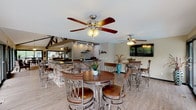 Located at 1004 La Bonne Pkwy in Manchester, Missouri, this single-story community clubhouse offers a well-appointed gathering space designed for both relaxation and socializing. The main lounge area is an architectural standout, featuring soaring vaulted ceilings with dramatic exposed dark steel beams and trusses that draw the eye upward. Abundant natural light pours in through floor-to-ceiling windows and glass doors lining the perimeter, offering unobstructed views of the surrounding greenery and the outdoor deck beyond. The lounge is furnished with plush, inviting sofas and accent chairs arranged around a central coffee table, creating a warm and comfortable atmosphere. Light hardwood flooring flows throughout, complementing the neutral warm-toned walls. Adjacent to the lounge, a dedicated bar and dining area features a stacked-stone accent bar with dark countertops, track lighting, and pub-height tables with cushioned seating — accented by a playful "Cheers" wall sign. A sunroom-style dining nook with vaulted ceilings, large sliding glass doors, and carpet flooring provides a quieter setting with deck access and views of the surrounding community. Ceiling fans are installed throughout for year-round comfort. The deck, accessible from multiple points, offers outdoor seating surrounded by mature trees, enhancing the overall resort-like ambiance of this inviting clubhouse.