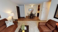 This single-floor unit presents an open, flowing layout that connects the living, dining, and kitchen areas into one cohesive space. The living area is finished with plush carpeting and is furnished with comfortable seating arranged around a dark wood coffee table, creating a relaxed and inviting atmosphere. A decorative plant adds a touch of greenery to the space.

Transitioning toward the dining area, the flooring shifts to a warm hardwood-style surface that extends through to the kitchen. The dining space accommodates a round, dark-finished table with matching chairs, ideal for everyday meals or casual gatherings. A framed piece of wall art and a ceiling light fixture above the table add subtle character to the area.

Adjacent to the dining space, the kitchen is partially visible and features dark cabinetry with lighter countertops, providing a clean, functional workspace. A coat closet and entry door are positioned nearby, offering convenient access and practical storage.

Throughout the unit, neutral wall tones and consistent finishes contribute to a cohesive, understated aesthetic, while the open sightlines between rooms enhance the overall sense of space and connectivity.