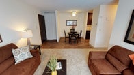 This single-floor unit presents an open, flowing layout that connects the living, dining, and kitchen areas into one cohesive space. The living area is finished with plush carpeting and is furnished with comfortable seating arranged around a dark wood coffee table, creating a relaxed and inviting atmosphere. A decorative plant adds a touch of greenery to the space.

Transitioning toward the dining area, the flooring shifts to a warm hardwood-style surface that extends through to the kitchen. The dining space accommodates a round, dark-finished table with matching chairs, ideal for everyday meals or casual gatherings. A framed piece of wall art and a ceiling light fixture above the table add subtle character to the area.

Adjacent to the dining space, the kitchen is partially visible and features dark cabinetry with lighter countertops, providing a clean, functional workspace. A coat closet and entry door are positioned nearby, offering convenient access and practical storage.

Throughout the unit, neutral wall tones and consistent finishes contribute to a cohesive, understated aesthetic, while the open sightlines between rooms enhance the overall sense of space and connectivity.