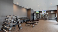 Located at 1206 N. 4th Street in Columbus, Ohio, this professionally designed fitness center offers a generous, single-floor layout that accommodates a wide range of workout needs. The space is divided into well-organized zones, each dedicated to a specific type of training, creating an efficient and focused exercise environment.

The cardio area is anchored by a row of treadmills and stationary bikes, positioned alongside floor-to-ceiling glass windows framed in dark steel that flood the space with natural light and provide views to the outdoors. A striking sputnik-style chandelier accents this section, adding a refined architectural touch.

The strength training zone is extensively equipped, featuring a full rack of dumbbells ranging from lighter to heavy weights, multiple adjustable benches, cable machines, and a variety of selectorized strength machines. Wall-mounted flat-screen TVs are positioned throughout for entertainment during workouts. Large mirrors reflect the open layout and contribute to the sense of spaciousness.

An additional functional training area includes a heavy punching bag with TRX suspension straps and open floor space for dynamic exercises. A dedicated amenity station near the entrance provides cleaning supplies and storage. The overall aesthetic is sleek and contemporary, with dark gray accent walls, recessed lighting, and durable rubber and carpet flooring throughout.