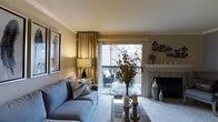 This single-floor living space offers a thoughtfully designed living room that balances comfort and style. The room is anchored by a tiled fireplace, which serves as a focal point along one wall, flanked by decorative ceramic vases that add an artistic touch. Crown molding along the ceiling lends a refined architectural detail to the space.

Natural light fills the room through a large sliding glass door dressed with elegant gold drape panels, which opens directly onto a private balcony. The outdoor space is visible through the glass, featuring seating and greenery, creating a seamless indoor-outdoor connection. A plush, carpeted floor complements the soft grey sofa, which is styled with nature-inspired throw pillows.

The living area is further enhanced by a mirrored coffee table styled with candles and a striking floral arrangement in a glass vase, adding warmth and texture. A framed artwork piece on the wall and a classic table lamp contribute to the inviting ambiance. Overall, the space presents a warm, well-appointed atmosphere suited for comfortable everyday living.