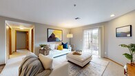 This single-floor residence offers a thoughtfully arranged layout that balances open communal spaces with comfortable private retreats. The living room serves as the heart of the home, featuring neutral gray walls, soft carpeting, and large French doors that invite abundant natural light and provide direct access to an outdoor area. A bold abstract artwork anchors the main wall, complementing the contemporary furnishings and layered area rug beneath.

The living space flows seamlessly into a dedicated dining area, defined by wood-look flooring that transitions from the carpeted lounge zone. A compact, well-equipped galley kitchen runs parallel to both walls, featuring warm honey-toned cabinetry, white countertops, recessed lighting, and full appliances including a dishwasher, range, and refrigerator.

A hallway connects the main living areas to two bedrooms, each carpeted and bathed in natural light from generously sized windows. The primary bedroom includes both a walk-in closet and an en-suite bathroom, while a second full bathroom with a tub-shower combination serves the remainder of the residence. Both bathrooms feature wood-look flooring and warm wood vanity cabinetry, maintaining a consistent aesthetic throughout the home.